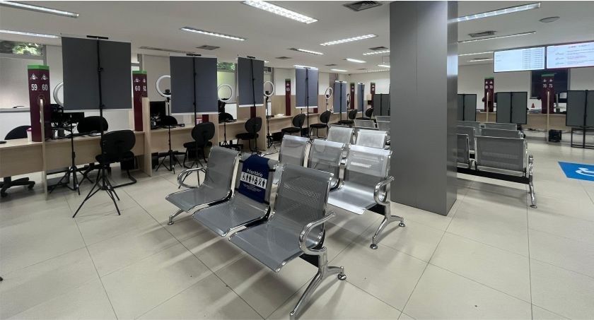 Foto mostra guichês de atendimento e cadeiras de espera em uma sala com piso e parede claros.