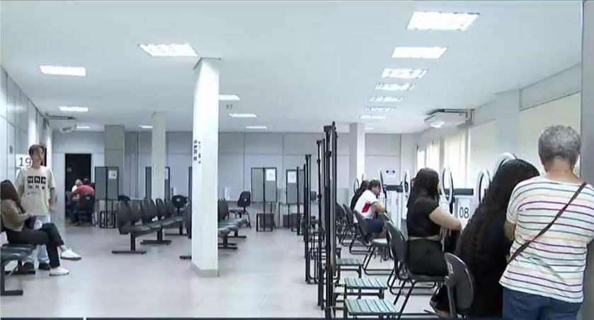 Foto mostra pessoas sendo atendidas e aguardando atendimento em uma sala com vários guichês e ca...