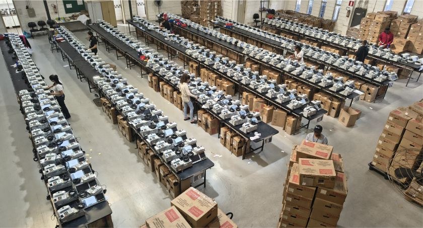 Foto mostra dezenas de urnas eletrônicas sobre bancadas.