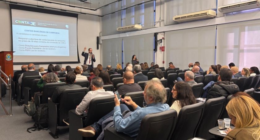Foto do auditório do TRE-MG. Em primeiro plano, parte da plateia. Ao fundo, um homem de pé com o...