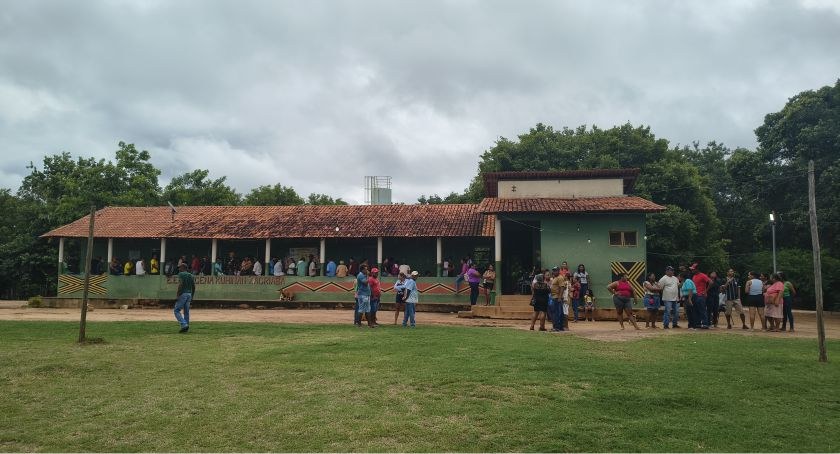 Foto mostra escola em aldeia indígena. Várias pessoas estão em fila na varanda da escola.