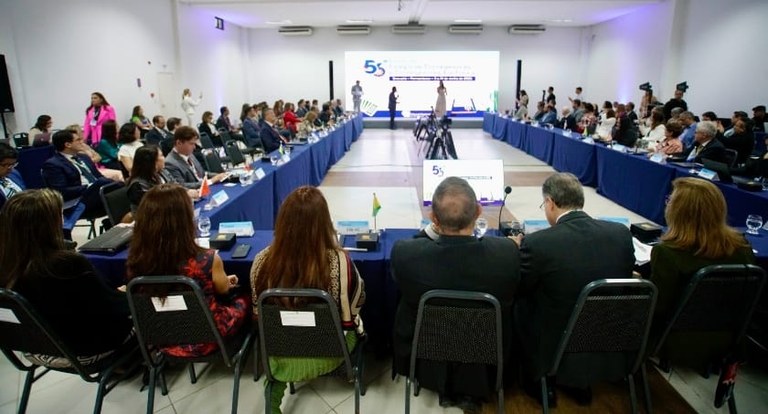 Diversos homens e mulheres sentados ao redor de uma mesa em formato de U. Ao fundo, um homem e u...