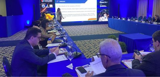 Foto mostra alguns homens e mulheres sentados ao redor de uma mesa em formato de U. A diretora-g...