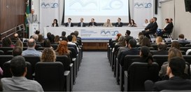 Foto mostra a plateia de um auditório em primeiro plano, com várias pessoas sentadas. Ao fundo, ...