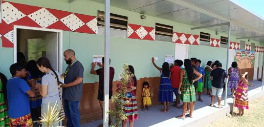 Diversos indígenas observam cartazes colados na parede de uma escola.