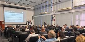 Foto do auditório do TRE-MG. Em primeiro plano, parte da plateia. Ao fundo, um homem de pé com o...