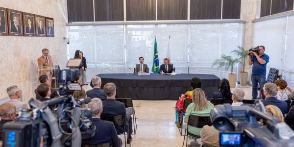 Ao fundo, presidente do TRE-MG e ALMG sentados à mesa. No primeiro plano, plateia sentada de cos...