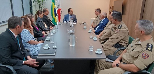 Assinatura de convênio TRE-MG e PMMG Representantes do TRE-MG e da Polícia Militar sentados ao redor de uma mesa retangular. Ao fundo...