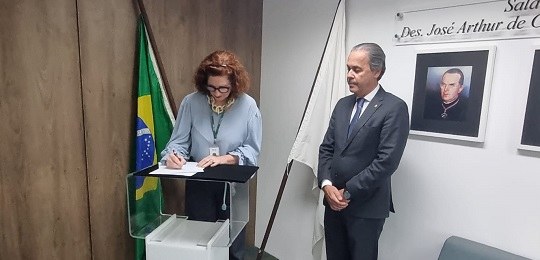 Desembargador Maurício Soares observa Elizabeth Jucá assinando um documento. Os dois estão de pé...