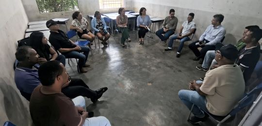 Foto mostra várias pessoas sentadas em formato de círculo, dentro de uma sala de aula.