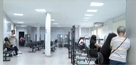Foto mostra pessoas sendo atendidas e aguardando atendimento em um ambiente com vários guichês e...