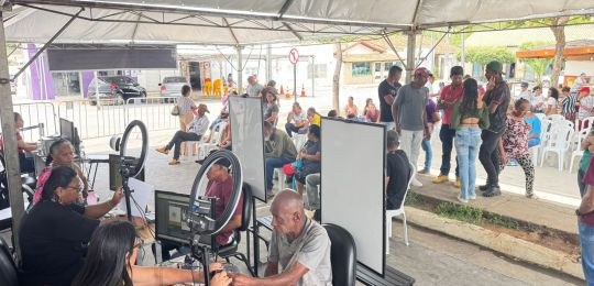 Foto mostra diversas pessoas sentadas em cadeiras debaixo de uma tenda branca. No canto direito,...
