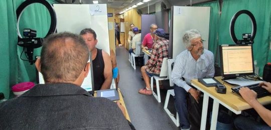 Foto mostra diversas pessoas sentadas em cadeiras debaixo de uma tenda branca. No canto direito,...