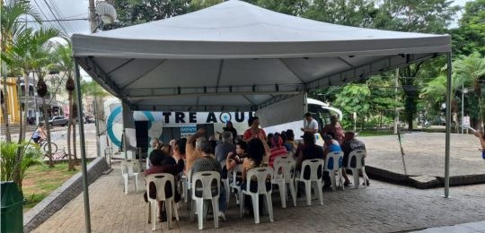 Foto mostra diversas pessoas sentadas em cadeiras debaixo de uma tenda branca. No canto direito,...