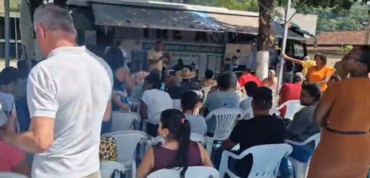 Foto mostra diversas pessoas sentadas em cadeiras debaixo de uma tenda branca. No canto direito,...