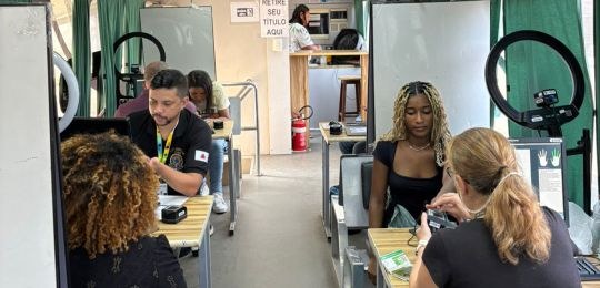 Foto mostra diversas pessoas sentadas em cadeiras debaixo de uma tenda branca. No canto direito,...