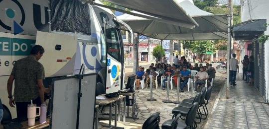 Foto mostra diversas pessoas sentadas em cadeiras debaixo de uma tenda branca. No canto direito,...