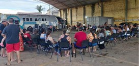Foto mostra várias pessoas sentadas em cadeiras em um galpão. Ao fundo, um ônibus branco com a i...