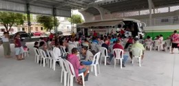 Foto mostra dezenas de pessoas sentadas em cadeiras brancas em um galpão. Ao fundo, um ônibus br...