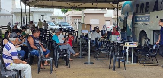 Foto mostra diversas pessoas sentadas em cadeiras debaixo de uma tenda branca. No canto direito,...