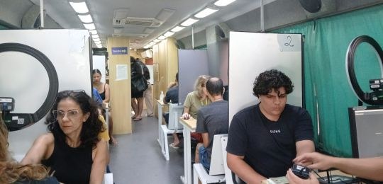 Foto mostra diversas pessoas sentadas em cadeiras debaixo de uma tenda branca. No canto direito,...