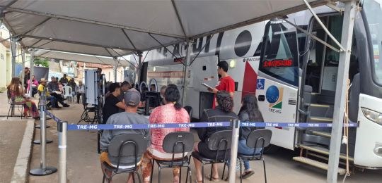 Foto mostra eleitoras e eleitores sentados do lado de fora de um ônibus que tem a marca TRE Aqui...