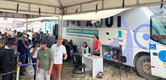 Foto mostra diversas pessoas sentadas em cadeiras debaixo de uma tenda branca. No canto direito,...
