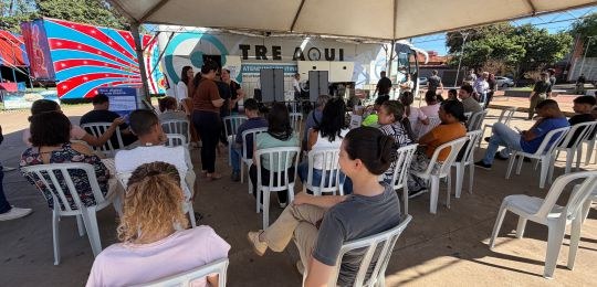 Foto mostra diversas pessoas sentadas em cadeiras debaixo de uma tenda branca. No canto direito,...