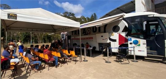 Foto mostra pessoas aguardando atendimento debaixo de uma tenda branca, em frente ao ônibus TRE ...