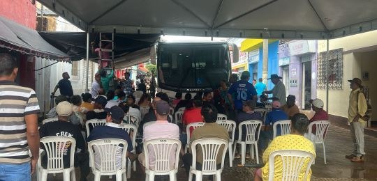 Foto mostra diversas pessoas sentadas em cadeiras debaixo de uma tenda branca. No canto direito,...