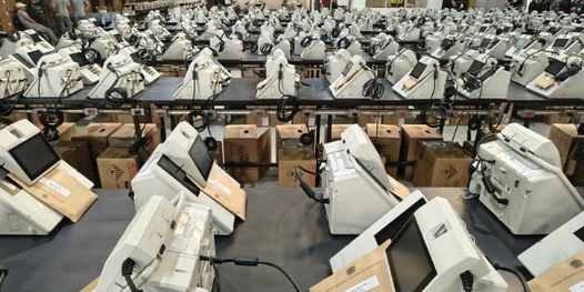 Dezenas de urnas eletrônicas sobre bancadas. Em primeiro plano, é possível ver que há um envelop...