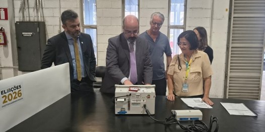Dezenas de urnas eletrônicas sobre bancadas. Em primeiro plano, é possível ver que há um envelop...