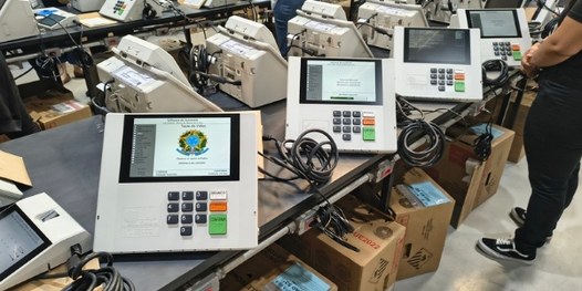 Dezenas de urnas eletrônicas sobre bancadas. Em primeiro plano, é possível ver que há um envelop...
