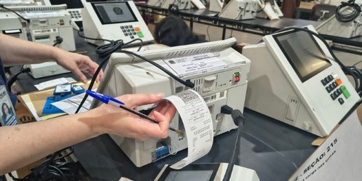 Dezenas de urnas eletrônicas sobre bancadas. Em primeiro plano, é possível ver que há um envelop...