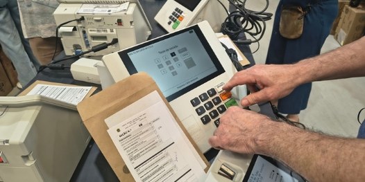 Dezenas de urnas eletrônicas sobre bancadas. Em primeiro plano, é possível ver que há um envelop...