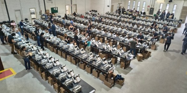 Foto tirada de cima mostra dezenas de urnas eletrônicas sobre bancadas de madeira. Algumas pesso...