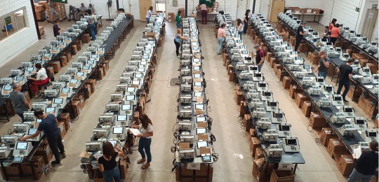 Carga das urnas BH 2º turno 2024 Foto mostra dezenas de urnas eletrônicas sobre bancadas. Algumas pessoas estão de pé perto das b...