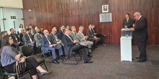 Foto de um pequeno auditório. No canto direito, desembargador Nicolau Lupianhes de pé. No canto ...