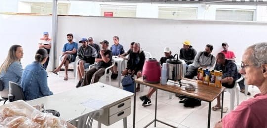 Foto mostra duas servidoras do TRE-MG conversando com alguns homens. Todos estão sentados em um ...