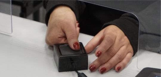 Foto mostra as mãos de uma mulher morena com as unhas pintadas de vermelho. O polegar da mão dir...