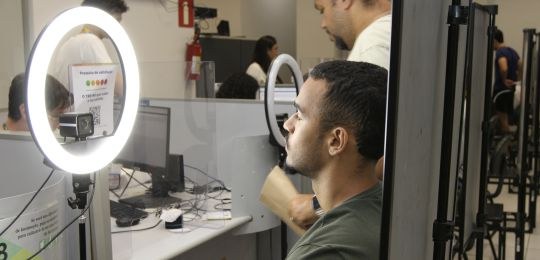 Foto mostra um jovem branco de cabelo curto sentado junto a um painel branco.