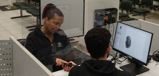 Rapaz observa uma mulher assinando em equipamento digital, assinatura aparece na tela do computa...