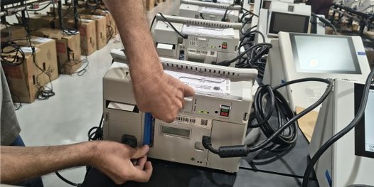 Foto mostra dezenas de urnas eletrônicas sobre bancadas, dentro de um galpão. Algumas pessoas es...
