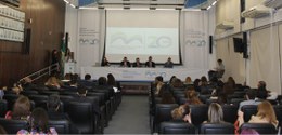 Foto mostra a plateia de um auditório em primeiro plano e cinco homens sentados junto a uma mesa...