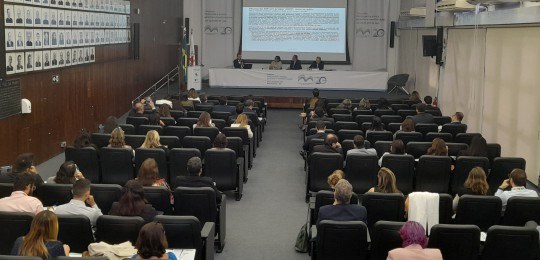 Foto mostra a plateia de um auditório em primeiro plano e quatro homens sentados junto a uma mes...