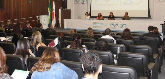 Foto mostra parte da plateia de um auditório. Ao fundo, quatro mulheres sentadas junto a uma mes...