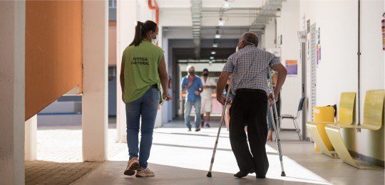 Foto de uma mulher jovem com colete verde ao lado de um homem idoso andando com muletas.