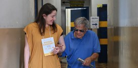 Duas pessoas com os braços dados, do lado esquerdo uma mulher de roupa bege auxilia um idoso de ...