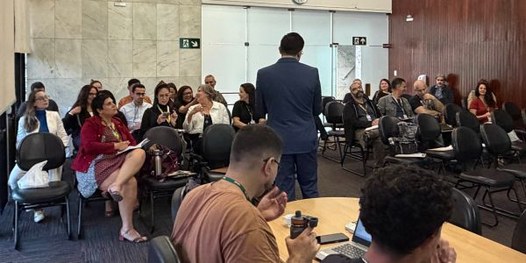 Foto mostra um homem de pé, de costas para a câmera. Ele fala para homens e mulheres sentados em...
