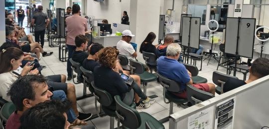 Foto mostra várias pessoas sentadas e alguns guichês de atendimento.